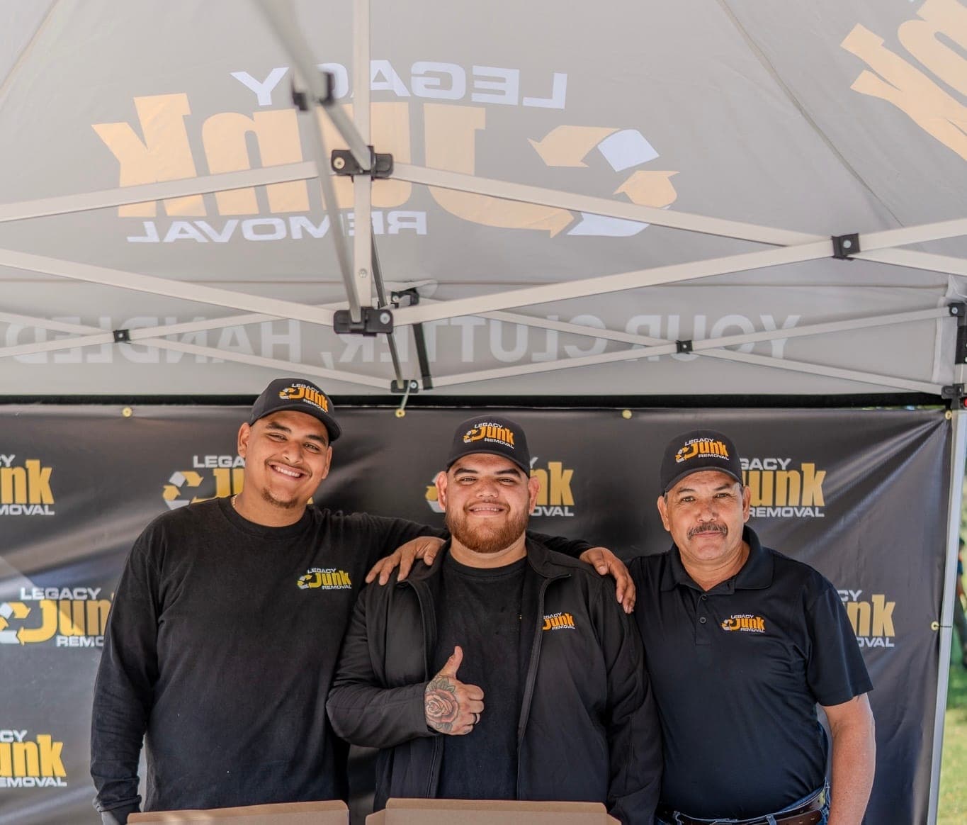 Three members of the Legacy crew in black Legacy shirts and caps smiling in front of the Legacy Junk Removal backdrop